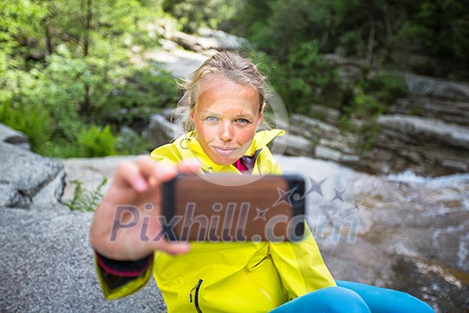 Pretty, young female hiker taking a selfie while outdoors on a hike, taking a break near a mountain stream (shallow DOF)