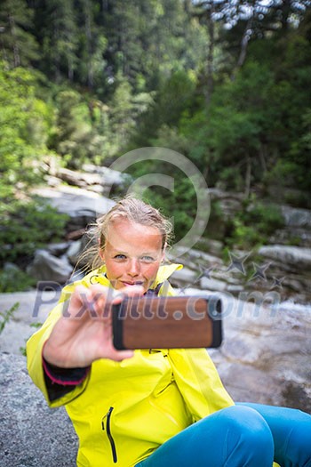 Pretty, young female hiker taking a selfie while outdoors on a hike, taking a break near a mountain stream (shallow DOF)