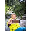 Pretty, young female hiker taking a selfie while outdoors on a hike, taking a break near a mountain stream (shallow DOF)