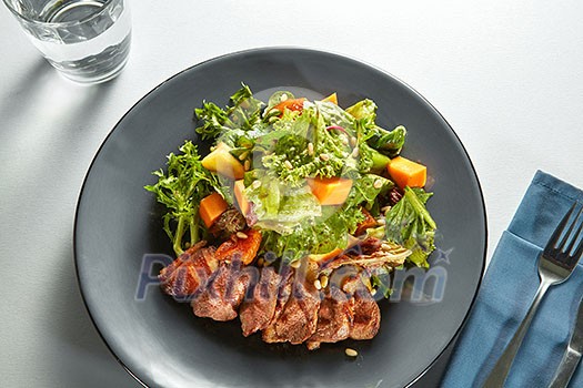 grilled duck meat with roasted pumpkin and fresh green salad on a plate with cutlery on a table