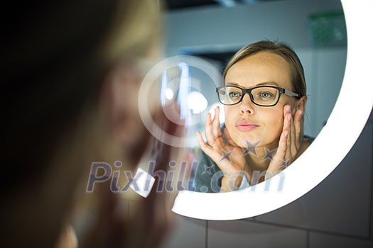 Pretty, young woman in front of her bathroom mirror in the morning - checking her face, getting ready for work (color toned image; shallow DOF) Pretty, young woman in front of her bathroom mirror in the morning - checking her face, getting ready for work (color toned image; shallow DOF)