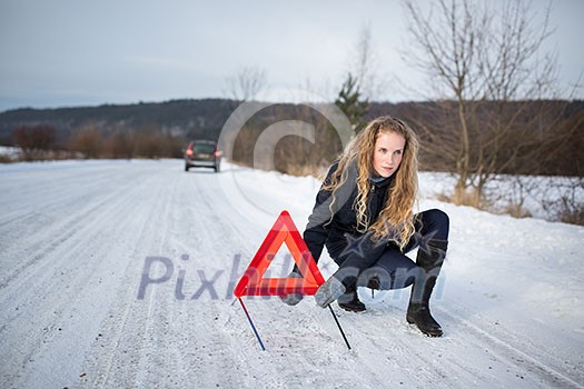 Young woman setting up a warning triangle and calling for assistance after her car broke down in the middle of nowhere on a freezing winter day