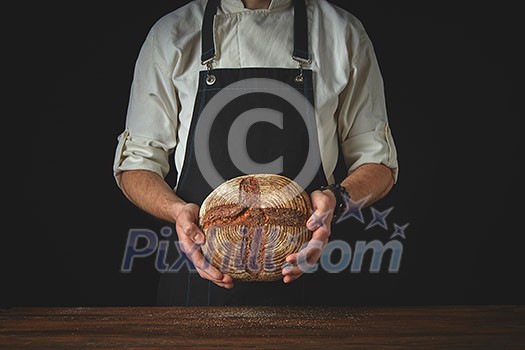 baker in an apron holds bread in hands on a black background