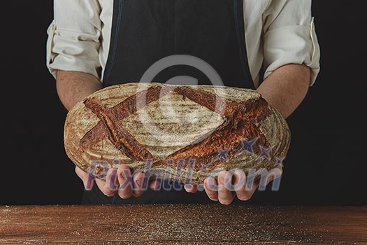 On the black background of a wooden table the baker's hands hold fresh organic oval bread On the black background of a wooden table the baker's hands hold fresh organic oval bread