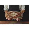 On the black background of a wooden table the baker's hands hold fresh organic oval bread