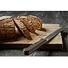 Loaf of sliced whole grain bread on wood bread board with knife, on a dark background