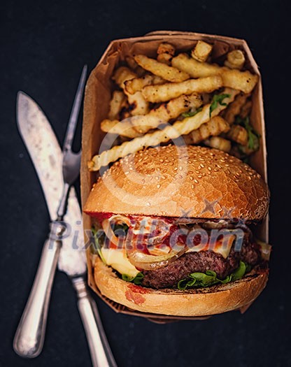 Fresh homemade hamburger with fried potatoes packed in a box on a black background. Top view. Retro vintage toned. Fresh homemade hamburger with fried potatoes packed in a box on a black background. Top view. Retro vintage toned.