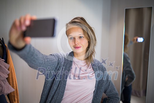 Young woman shopping in a fashion store, trying on some clothes she likes  - taking a selfie picture with her smartphone in the cabin (color toned image; shallow DOF)