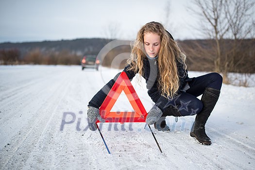 Young woman setting up a warning triangle and calling for assistance after her car broke down in the middle of nowhere on a freezing winter day