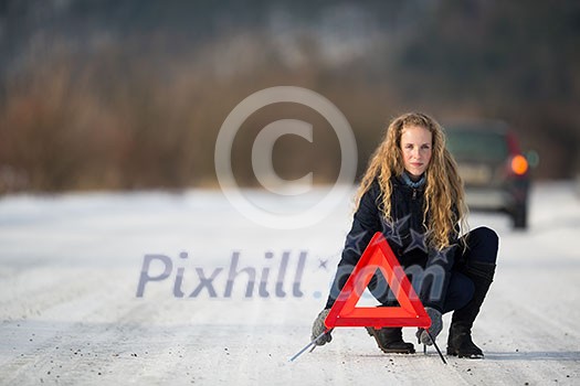 Young woman setting up a warning triangle and calling for assistance after her car broke down in the middle of nowhere on a freezing winter day