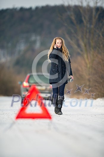 Young woman setting up a warning triangle and calling for assistance after her car broke down in the middle of nowhere on a freezing winter day