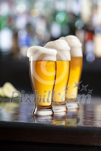 three glasses of light beer with foam served on wooden desk. Bar on background three glasses of light beer with foam served on wooden desk. Bar on background