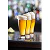 three glasses of light beer with foam served on wooden desk. Bar on background