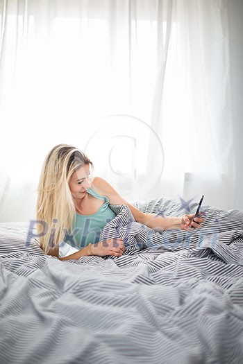 Beautiful young woman in bed, taking a selfie on her cell phone (color toned image; shallow DOF)