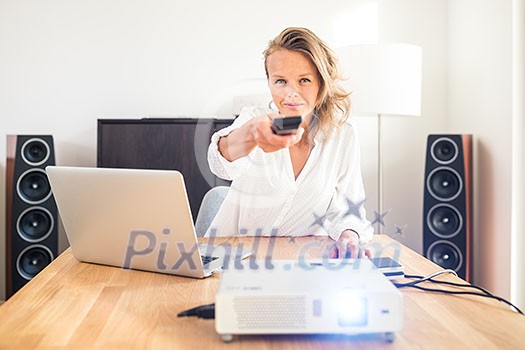 Pretty, female freelancer working from home, preparing led projector for a client presentation - using remote control for projector Pretty, female freelancer working from home, preparing led projector for a client presentation - using remote control for projector