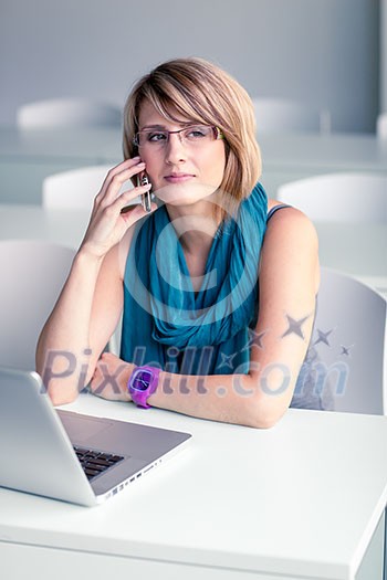 Pretty, young woman at an office, using a laptop and her smartphone