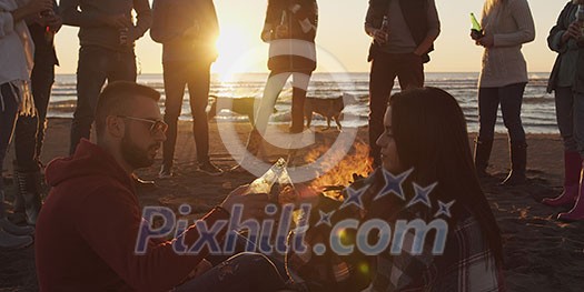 Happy Carefree Young Friends Having Fun And Drinking Beer By Bonefire On The Beach As The Sun Begins To Set