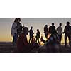 Happy Carefree Young Friends Having Fun And Drinking Beer By Bonefire On The Beach As The Sun Begins To Set