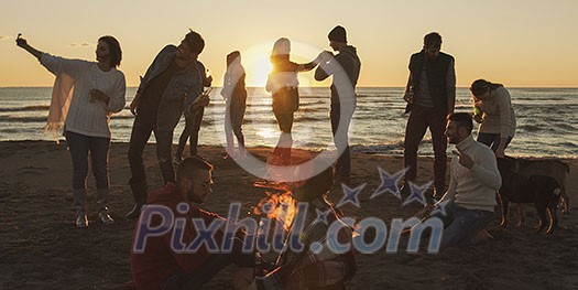 Happy Carefree Young Friends Having Fun And Drinking Beer By Bonefire On The Beach As The Sun Begins To Set