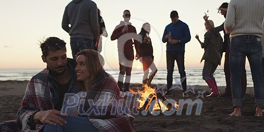 Happy Carefree Young Friends Having Fun And Drinking Beer By Bonefire On The Beach As The Sun Begins To Set