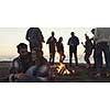 Happy Carefree Young Friends Having Fun And Drinking Beer By Bonefire On The Beach As The Sun Begins To Set