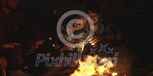 Young Couple Sitting with friends Around Campfire on The Beach At Night drinking beer
