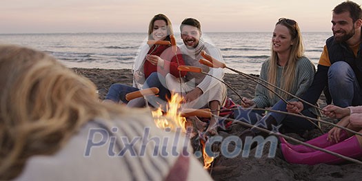 Group of young friends sitting by the fire late at night, grilling sausages and drinking beer, talking and having fun