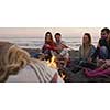 Group of young friends sitting by the fire late at night, grilling sausages and drinking beer, talking and having fun