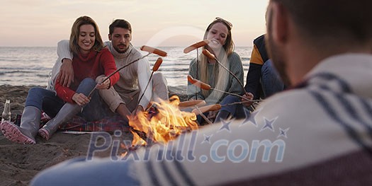 Group of young friends sitting by the fire late at night, grilling sausages and drinking beer, talking and having fun Group of young friends sitting by the fire late at night, grilling sausages and drinking beer, talking and having fun