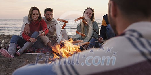 Group of young friends sitting by the fire late at night, grilling sausages and drinking beer, talking and having fun