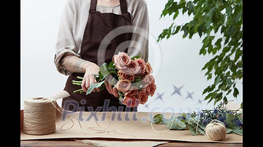 A woman florist makes a bouquet of beige roses cappuccino and blue eryngiums, wraps flowers in paper and tie a string on a wooden table. Flower Shop, Video footage 1080p full HD