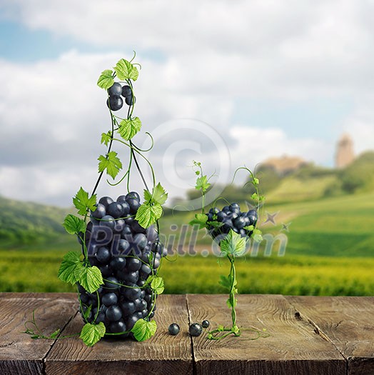 A glass and a bottle of wine made by of grape leaves and a bunch of grapes on a wooden background with a blurred background overlooking the green peaks