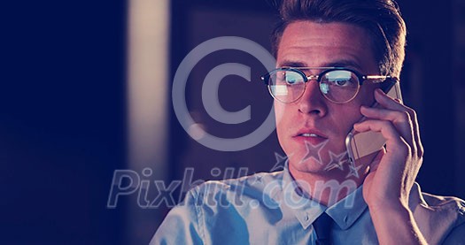 Young man using mobile phone while working on computer at night in dark office.