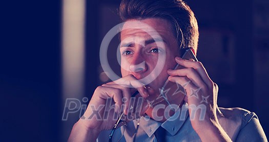 Young man using mobile phone while working on computer at night in dark office.