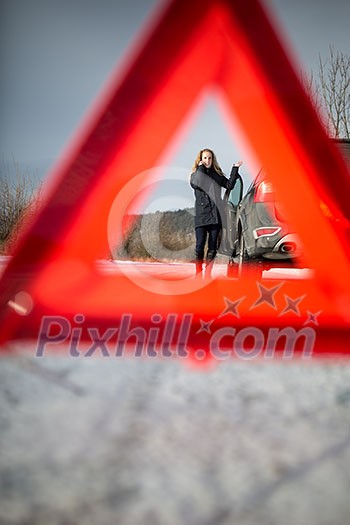 Realy angry young woman in a road distress situation - setting up a warning triangle and calling for assistance after her car broke down in the middle of nowhere on a freezing winter day