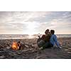 Young Couple Relaxing By The Fire, Drinking A Beer Or A Drink From The Bottle on the beach at autumn day