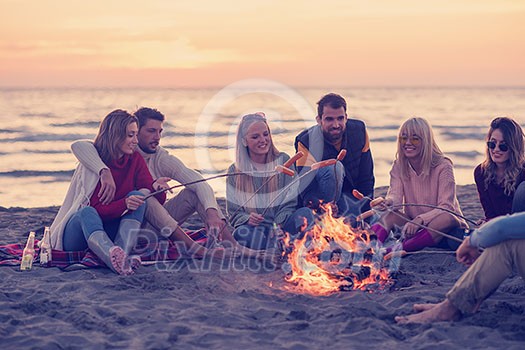 Group of young friends sitting by the fire at autumn beach, grilling sausages and drinking beer, talking and having fun