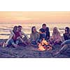 Group of young friends sitting by the fire at autumn beach, grilling sausages and drinking beer, talking and having fun