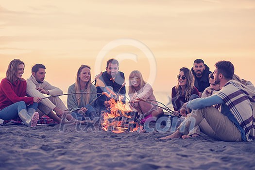 Group of young friends sitting by the fire at autumn beach, grilling sausages and drinking beer, talking and having fun