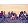 Group of young friends sitting by the fire at autumn beach, grilling sausages and drinking beer, talking and having fun