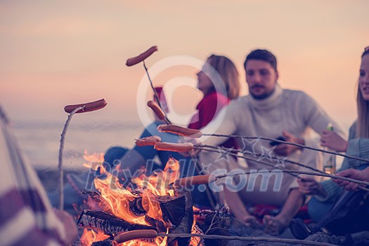 Group of young friends sitting by the fire at autumn beach, grilling sausages and drinking beer, talking and having fun Group of young friends sitting by the fire at autumn beach, grilling sausages and drinking beer, talking and having fun