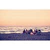 Group of young friends sitting by the fire at autumn beach, grilling sausages and drinking beer, talking and having fun