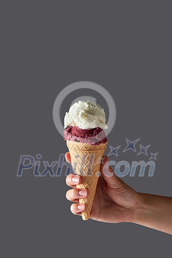 A red fruit ball and a vanilla white ice cream ball in a waffle cone held by a woman's hand against a gray background. Summer concept with copy space. A red fruit ball and a vanilla white ice cream ball in a waffle cone held by a woman's hand against a gray background. Summer concept with copy space.