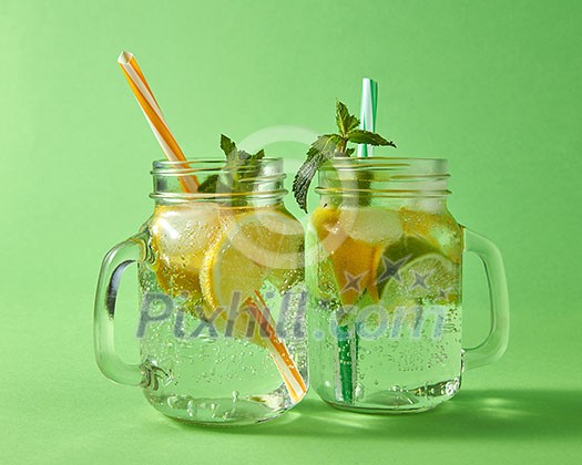 Cold natural lemonade handmade with bubbles of air. Citrus fruits slices of lemon and lime, ice, leaf of mint, water with plastic straws in the glass. Two glass jar on green background.