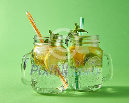 Cold natural lemonade handmade with bubbles of air. Citrus fruits slices of lemon and lime, ice, leaf of mint, water with plastic straws in the glass. Two glass jar on green background. Cold natural lemonade handmade with bubbles of air. Citrus fruits slices of lemon and lime, ice, leaf of mint, water with plastic straws in the glass. Two glass jar on green background.