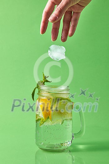 Glass jar on a green with homemade sparkling cocktail with slices of citrus fruits, green leaves of mint. A woman's hand throws a piece of ice into a glass jar. Glass jar on a green with homemade sparkling cocktail with slices of citrus fruits, green leaves of mint. A woman's hand throws a piece of ice into a glass jar.