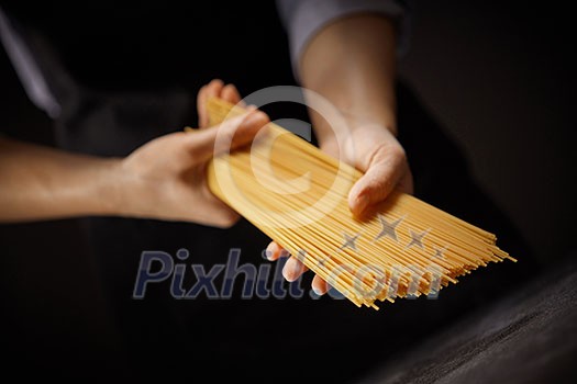 A cook in a black apron holds a raw long spaghetti on a dark background. Traditional Italian food A cook in a black apron holds a raw long spaghetti on a dark background. Traditional Italian food