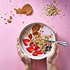 Healthy smoothie in white bowl with natural fruits, oat flakes and biscuits with woman's hands holding a bowl on pink background. Superfoods, natural detox, diet and healthy food. Top view