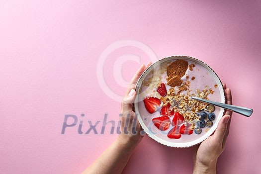 Healthy smoothie in white bowl with natural fruits, oat flakes and biscuits with woman's hands holding a bowl on pink background. Superfoods, natural detox, diet and healthy food. Flat lay