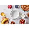 Empty plate, organic granola, banana, honey, milk, berries and almonds on a white wooden table with copy space. Ingredients for a healthy breakfast, step by step recipe. Top view